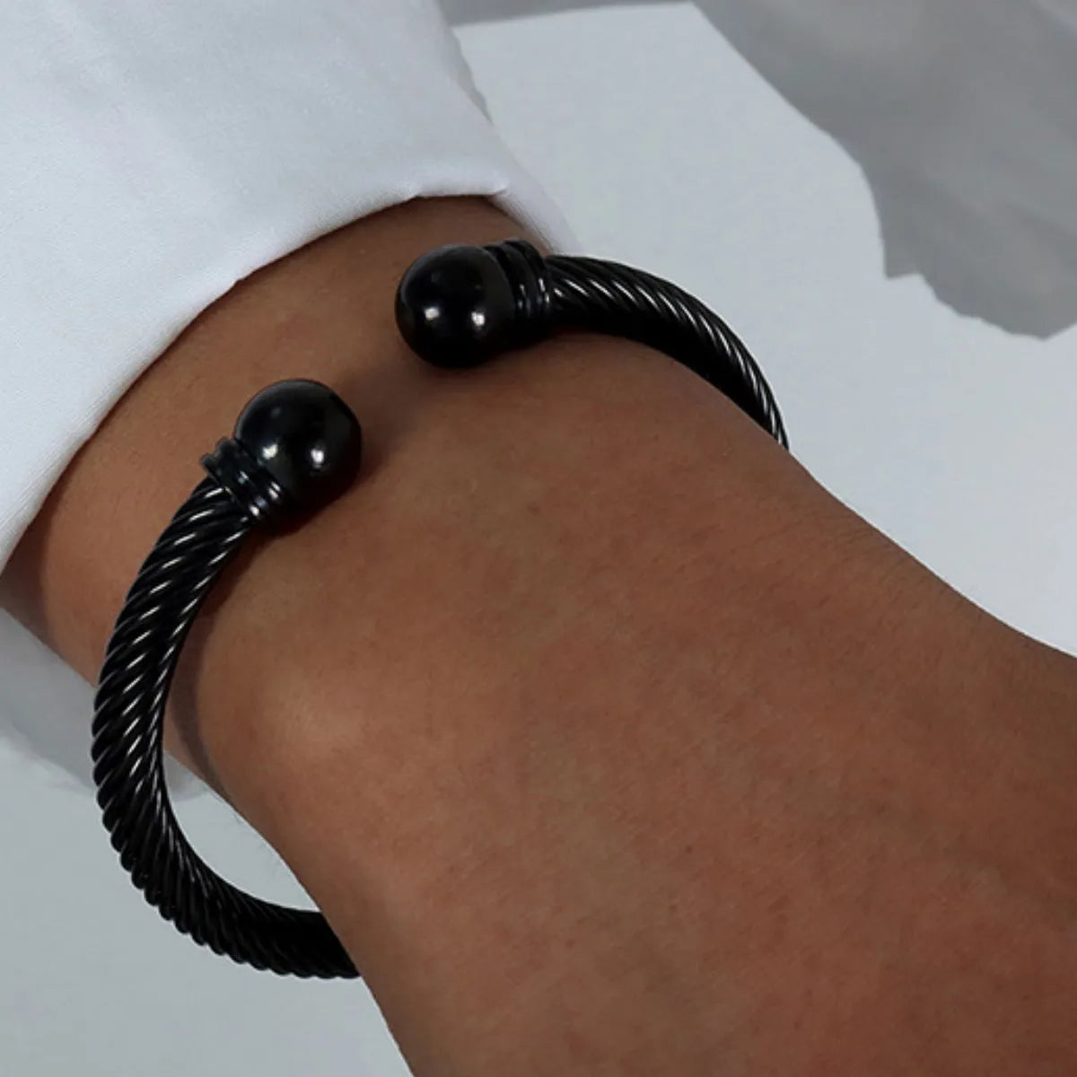 Black twisted cable cuff bracelet with spherical ends worn on wrist and white shirt sleeve visible