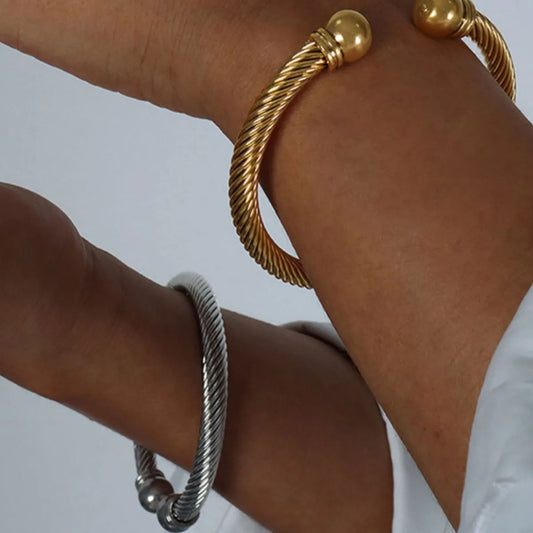 Close-up of two twisted cable cuff bracelets, one gold-plated and one silver, worn on wrists against a plain background.